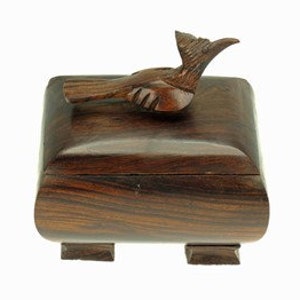 Peut inclure: Une boîte en bois foncé avec un oiseau sculpté sur le couvercle. La boîte a une forme carrée avec des bords arrondis et quatre petits pieds.