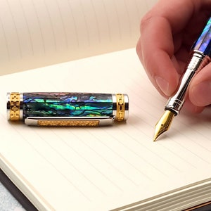 Op de afbeelding: Een gouden en zilveren vulpen met een blauw en groen abalone-schelplichaam. De pen ligt op een gelinieerd notitieboek.