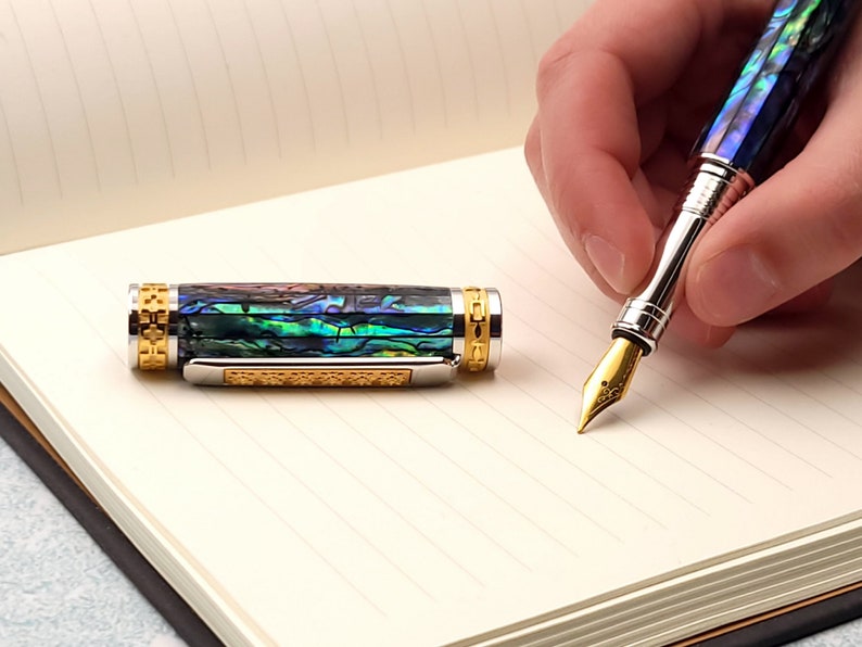 Op de afbeelding: Een gouden en zilveren vulpen met een blauw en groen abalone-schelplichaam. De pen ligt op een gelinieerd notitieboek.