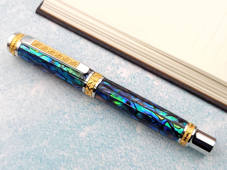 Op de afbeelding: Een blauwe en groene pen met gouden accenten en een zilveren dop. De pen is gemaakt van abalone-schelp en heeft een uniek, ingewikkeld ontwerp.