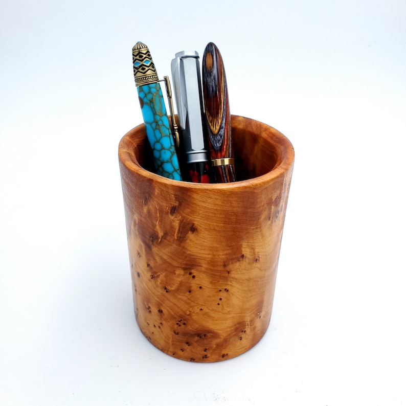 May include: A wooden pen holder with a natural wood grain finish. Three pens are inside the holder, one with a turquoise and gold design, one with a silver finish, and one with a red and black wood grain design.