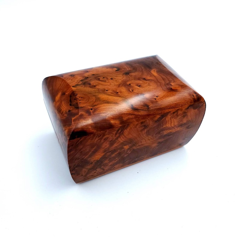 May include: A small, wooden box with a rich, reddish-brown color and a unique, swirling grain pattern. The box has a rounded, slightly curved top and a smooth, polished finish.