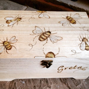 May include: A wooden box with a hinged lid and a brass latch. The box is decorated with a wood-burned design of bees and the name "Greta" is also wood-burned on the front.