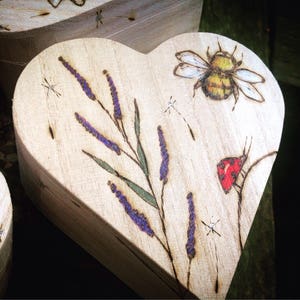 May include: A wooden heart-shaped box with a woodburned design of lavender flowers, a bee, and a ladybug. The box is a light brown color and the design is in shades of purple, yellow, and red.