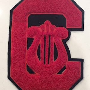 May include: A red felt letter "C" with a black border and a lyre design. The letter and lyre are raised and textured, with a black background. This patch is likely for a school or music-related organization.