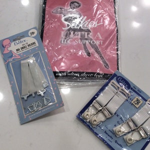 May include: Vintage lingerie accessories, including bra girdle holders and garter strips, are displayed. The items are packaged in blue and pink cardboard with the brand name "Beltx" and "Selkies Ultra TLC Support" printed on them. The garter strips are white.