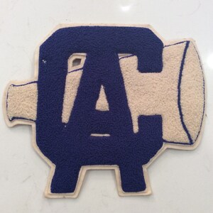 May include: A navy blue and cream-colored varsity letter patch. The letter "A" is prominently displayed, with a textured, fuzzy appearance. The patch has a unique shape, with a horn-like extension on the right side.
