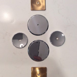 May include: An overhead shot of hardware components. The image features two square, gold-toned pieces with a centered square hole, and four circular, silver-toned pieces with a centered hole. The pieces are arranged in a cross-like pattern.