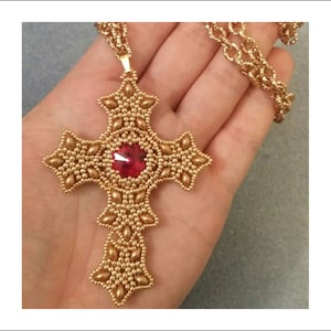 Può includere: Un ciondolo a forma di croce in perle dorate con una pietra preziosa rossa al centro. La croce è attaccata a una catena dorata.
