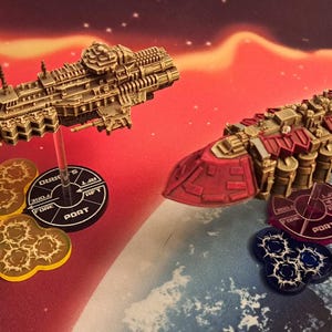 Puede incluir: Dos naves espaciales en miniatura, una dorada y otra roja, sobre bases circulares con marcas negras y blancas. Las bases están sobre una superficie azul y blanca con estrellas.