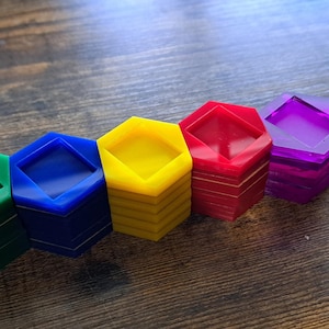 Può includere: Cinque pile di gettoni da gioco esagonali in plastica di colore verde, blu, giallo, rosso e viola. Ogni pila è composta da più pezzi identici con un centro quadrato incavato, disposti in fila su una superficie in legno marrone scuro.