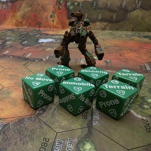 Op de afbeelding: Groene dobbelstenen met witte tekst, waaronder "Prone", "No Move", "Immobile", "Terrain" en "Skidding". De dobbelstenen liggen op een bruin en groen speelbord met een bruin en groen robotfiguur op de achtergrond.