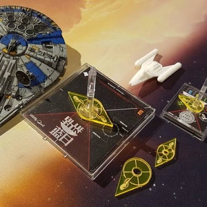 May include: A blue and grey spaceship miniature, a white spaceship miniature, two square black game bases with clear plastic flight stands, and three yellow translucent geometric game tokens. The larger base features white text including the characters 藍日.