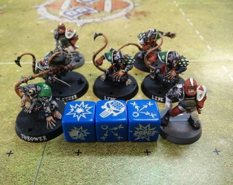 May include: A set of miniature figures for a tabletop game, featuring a team of rat-like creatures with tails and weapons. The figures are painted in green, black, and red, and are standing on black bases. There are three blue dice with white symbols, including a skull, a starburst, and arrows.