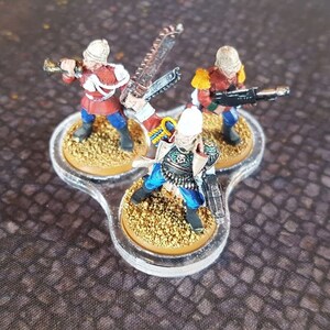 Może przedstawiać: Trzy miniaturowe figurki żołnierzy w czerwonych i niebieskich mundurach z białymi hełmami. Figurki stoją na przezroczystej plastikowej podstawie z trzema okrągłymi platformami. Figurki trzymają broń, w tym karabin, miecz i trąbę.
