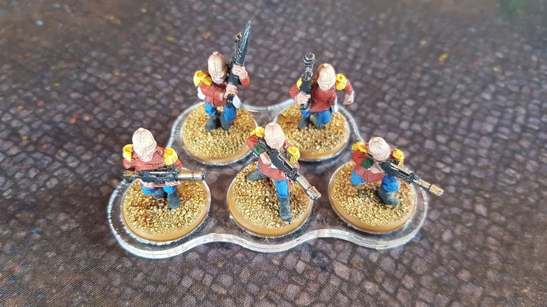 Może przedstawiać: Sześć miniaturowych figurek żołnierzy pomalowanych na czerwono, niebiesko i ż&oacute;łto, trzymających karabiny i stojących na okrągłych podstawach. Figurki są ułożone w k&oacute;łko na przezroczystej plastikowej podstawie.