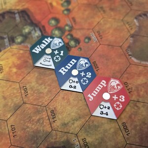 Puede incluir: Tres piezas de juego hexagonales con diferentes opciones de movimiento: "Caminar +1", "Correr +2" y "Saltar +3". Cada pieza tiene un c&iacute;rculo blanco en el centro y un escudo blanco con un n&uacute;mero en la parte inferior.