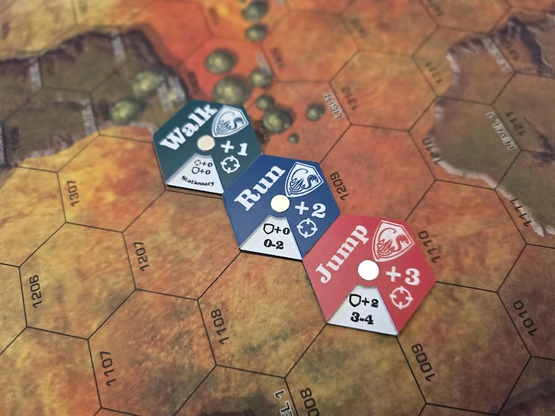 Puede incluir: Tres piezas de juego hexagonales con diferentes opciones de movimiento: "Caminar +1", "Correr +2" y "Saltar +3". Cada pieza tiene un c&iacute;rculo blanco en el centro y un escudo blanco con un n&uacute;mero en la parte inferior.