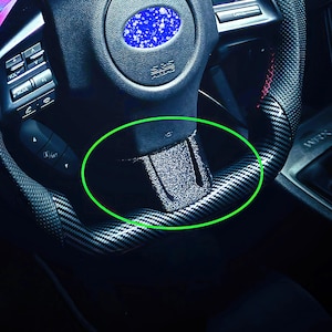 Puede incluir: Primer plano de un volante negro con un patrón de fibra de carbono y una tira reflectante plateada. El volante tiene un agarre de cuero negro y un botón plateado.