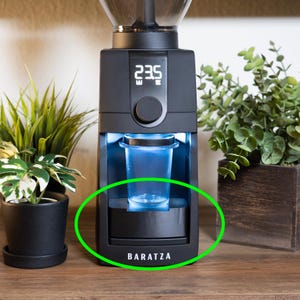 May include: A black Baratza coffee grinder with a digital display showing a number. A clear plastic cup is in place, ready to catch the ground coffee. The grinder is on a wooden surface, with plants in the background.