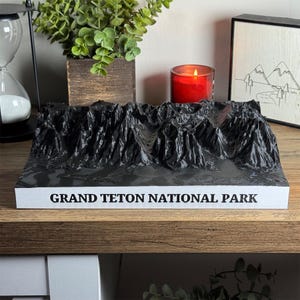 Puede incluir: Un modelo topográfico 3D del Parque Nacional Grand Teton. El modelo es negro y gris, con el nombre del parque impreso en blanco en la base. El modelo está sobre una estantería de madera con otros objetos decorativos.