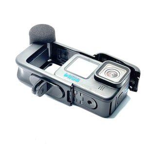 Op de afbeelding: Een zwarte actiecamera met een beschermend frame en een schuimrubberen windscherm. De camera heeft een lens, een scherm en de merknaam in blauw. Het frame heeft een bevestigingsmechanisme.