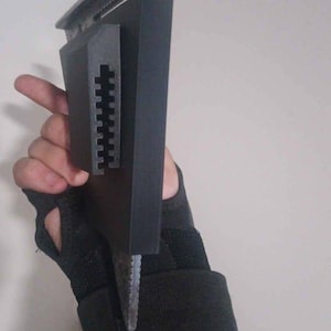 May include: A black and silver tool with a rectangular body and a metal blade. The tool has a textured grip and a silver top. The tool is held by a person wearing a black wrist support.