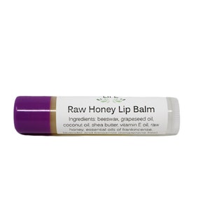 May include: A purple and beige lip balm tube with the text "Raw Honey Lip Balm" and a list of ingredients: beeswax, grapeseed oil, coconut oil, shea butter, vitamin E oil, raw honey, and essential oils of frankincense.