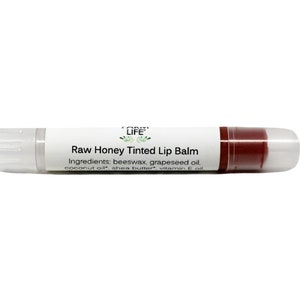 May include: A tube of Raw Honey Tinted Lip Balm. The lip balm is in a clear plastic tube with a white label. The label includes the product name, ingredients, and the brand logo. The lip balm itself is a reddish-brown color.