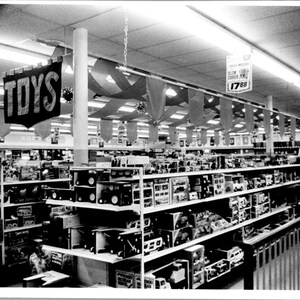 May include: Black and white photograph of a toy store interior. Shelves are filled with various toys, with a banner that reads "TOYS" hanging from the ceiling. The store is well-lit with overhead fluorescent lights.