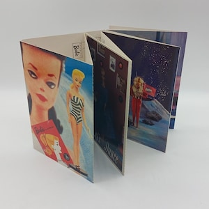 May include: A vintage Barbie book with a fold-out design. The cover features a close-up of Barbie's face. The inside pages feature images of Barbie in different outfits and settings, including a space scene with an astronaut Barbie.