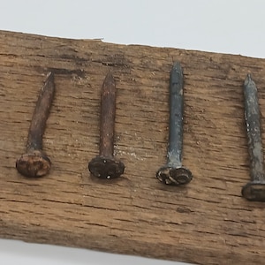 May include: Four antique nails with different finishes, including rusty, black, and silver, are displayed on a wooden surface. The nails have decorative heads with a raised design.
