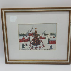 May include: Framed Christmas print featuring a festive village scene. The artwork depicts red houses, snow-covered ground, and a decorated Christmas tree. People are shown dancing around the tree. The frame is gold and white.
