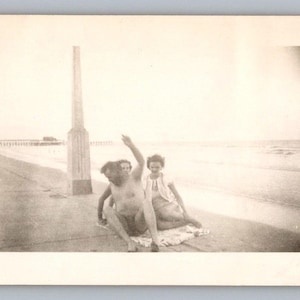 May include: Vintage black and white photograph of three people on a beach. One person is pointing upwards, while the other two are seated. A pier is visible in the background, and a tall wooden structure is on the left.