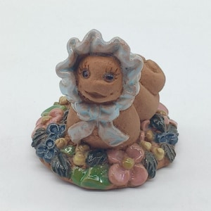 May include: A ceramic figurine of a smiling creature wearing a light blue bonnet with a matching bow. The figurine is brown and sits on a bed of colorful flowers, including pink, blue, and green.