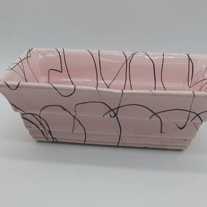 May include: A rectangular, pink ceramic planter with a black abstract design. The planter has a slightly flared shape and is suitable for indoor or outdoor use. The planter is approximately 10 inches long.