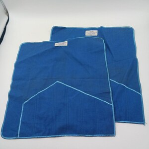 May include: Two blue cloths with light blue stitching and labels. The cloths are rectangular with a geometric design on one corner. The labels have text. The cloths are on a white background.