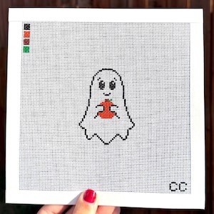 May include: A white cross-stitch canvas featuring a black-outlined ghost holding a red apple with a green leaf. The design includes a small color key in the top left corner with black, red, brown, and green squares, and the letters CC in the bottom right.