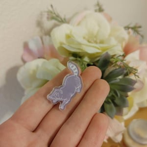 May include: A purple and white raccoon pin with a white outline. The pin is made of clear acrylic and has a shiny finish.