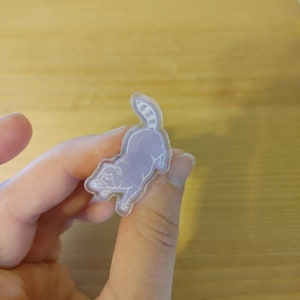 May include: A clear plastic pin with a white outline of a raccoon. The raccoon is facing to the left and has a striped tail.