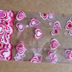 Puede incluir: Bolsa de regalo de plástico transparente con un patrón de corazones rosas y blancos. La bolsa tiene un borde festoneado y un patrón repetido de corazones.
