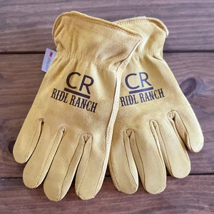 May include: A pair of tan leather work gloves with the initials "CR" and the text "RIDL RANCH" printed on each glove.