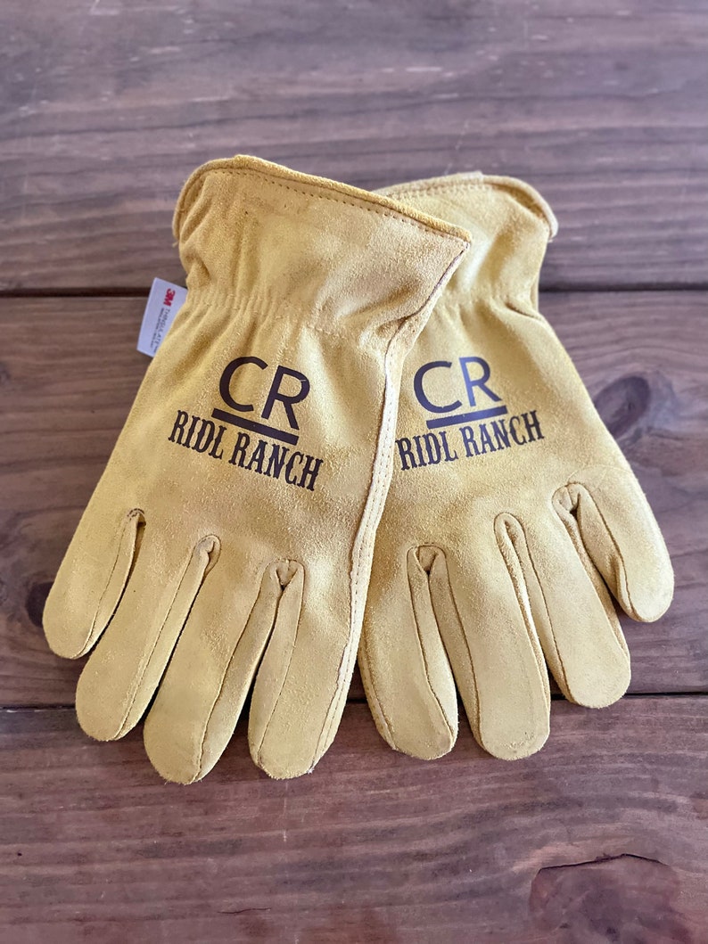May include: A pair of tan leather work gloves with the initials "CR" and the text "RIDL RANCH" printed on each glove.
