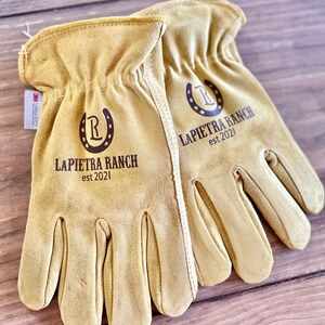 May include: A pair of tan leather work gloves with the text "LAPIETRA RANCH est 2021" and a horseshoe with the letter "R" inside printed on each glove.