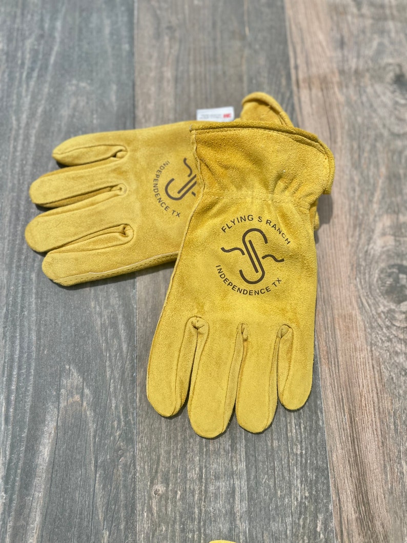 May include: A pair of yellow leather work gloves with the logo "Flying S Ranch Independence TX" printed on them.