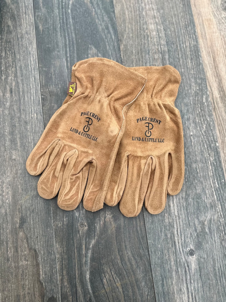 May include: A pair of brown leather work gloves with the text "PAGE CREST LAND & CATTLE LLC" and a logo of a "P" with a circle around it.