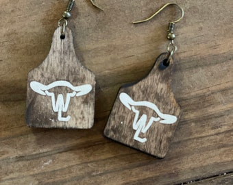 May include: A pair of wooden earrings with a cow brand design. The earrings are brown with a white cow brand design. The earrings are hanging from silver hooks.
