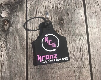 May include: A black key tag with a silver key ring. The tag has the text "KCG" inside a white circle with arrows, and the name "Kranz Custom Grinding" in pink and white.