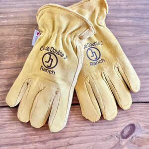 May include: A pair of tan leather work gloves with the text "Circle Double J Ranch" and a logo of a stylized "J" in a circle on each glove.