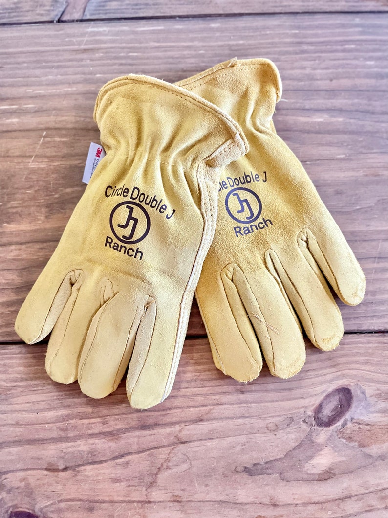 May include: A pair of tan leather work gloves with the text "Circle Double J Ranch" and a logo of a stylized "J" in a circle on each glove.